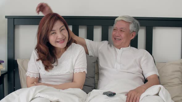 Asian elderly couple watching television enjoy love moment while lying on the bed. alt