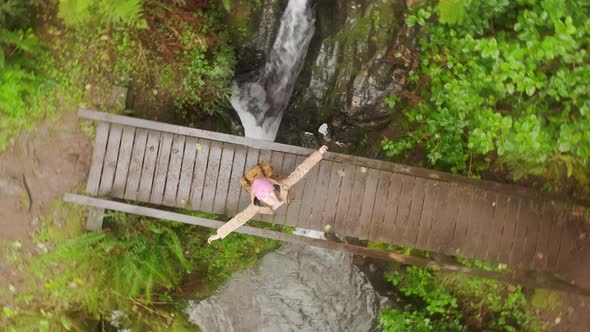 Happy Woman Tourist Spinning with Wide Opened Hands on Wooden Bridge Under Rain alt