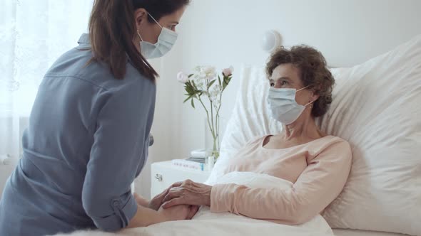 Home Caregiver With Face Mask Holding Hands of Female Senior Patient Lying in Bed at Nursing Home