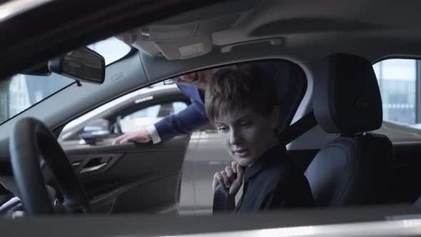Beautiful Young Girl with a Short Haircut Examines New Auto While Sitting in Passenger Compartment alt