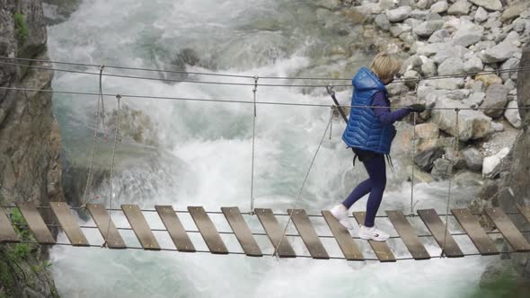 Female Traveler with a Backpack Walks Along an Old Wooden Bridge Over a Stormy Mountain River Aerial
