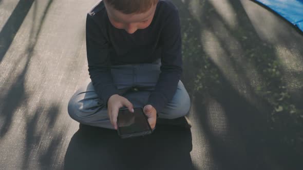 Boy Playing with Phone While Sitting on Trampoline