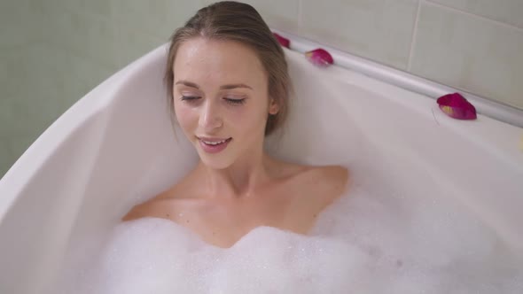 High Angle View Relaxed Young Slim Woman Lying in Bath Blowing Foam Smiling Looking at Camera alt