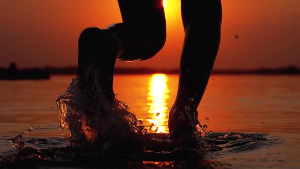 Silhouette of Legs of Boy Running Into the Water at Sunset and Creating Splashes. Slow Motion alt