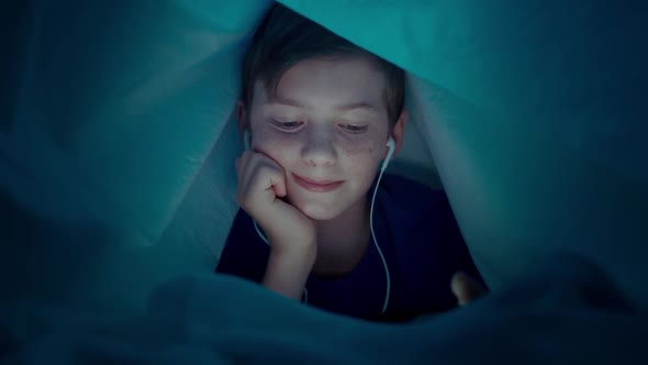 A Cute Boy with Headphones Makes a Video Call While Lying Under a Blanket at Night alt