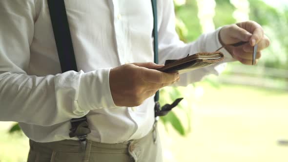 Wedding ceremony. Groom in suspenders opens leather notebook with vow and turn pages. Green plants alt