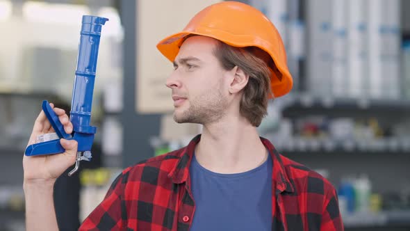 Closeup Portrait of Handsome Young Bearded Man in Hard Hat Posing with Caulking Gun in Hardware alt