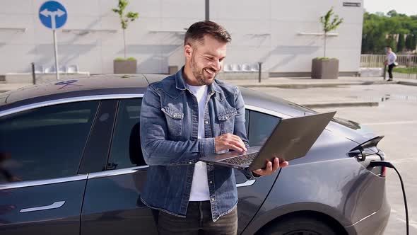 Man Enjoying Video Chat on Laptop while Waiting Until Refilling Stock of Energy on His Electric Car alt