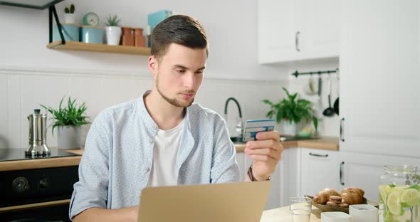 Man Pays Online By Card Using Laptop in Kitchen. Customer Makes Purchase at Home