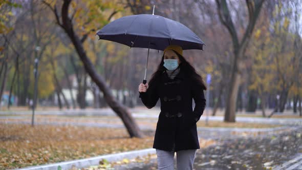 A Young Woman in a Protective Mask Walking in the Park Under Umbrella, Rainy Day, During Second Wave