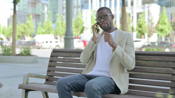 African Man Talking on Phone While Sitting Outdoor on Bench alt