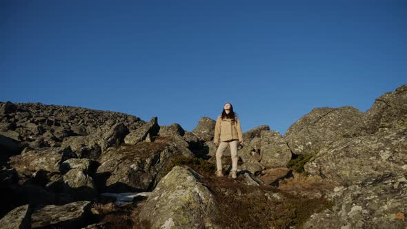 A Girl Traveler in a Beige Jacket Stands in the Mountains Raising Her Hands Up