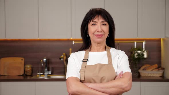 Portrait Smiling Aged Woman Chef Crosses Hands and Looks Camera in Home Kitchen alt