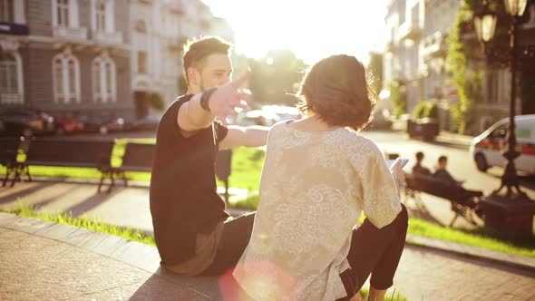 Young Beautiful Couple Smiling Speaking Looking at Tablet Sitting in City Park alt