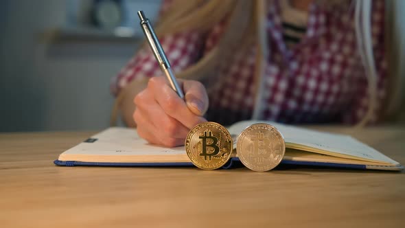 Woman Writing in Notebook with Bitcoins Nearby. Crop View of Female Hands in Checkered Shirt alt