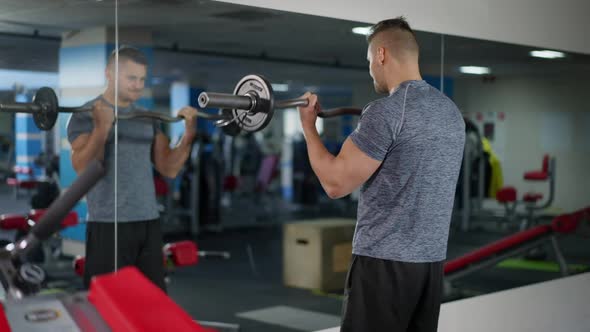 Back View of Strong Sportsman Lifting Heavy Barbell Reflecting in Mirror in Gym alt