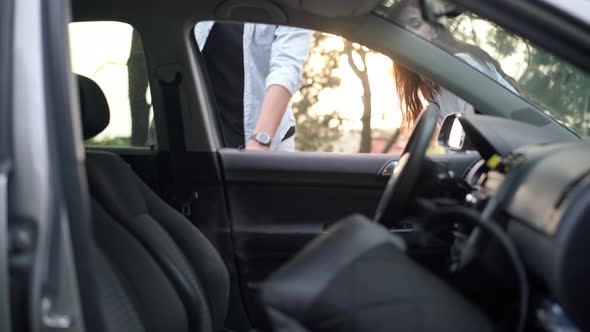 Young Man Sitting Down on Driver's Seat in Car with Backpack Talking to Joyful Woman alt