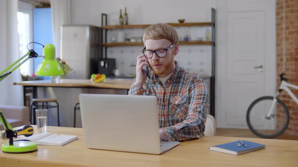 Freelancer Talking on Smartphone and Using Laptop at Home Office alt