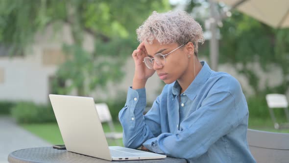 Young African Woman Thinking and Working on Laptop in Outdoor Cafe alt