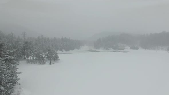 Drone flying over lake covered in white ice during snow storm towards forest alt