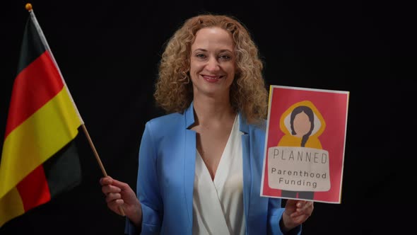 Smiling Confident Woman Holding German Flag and Planned Parenthood Funding Banner Looking at Camera alt