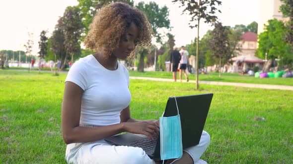 Young African American Woman Using Laptop in Park During Covid 19 Pandemic. Medical Disposable Mask alt
