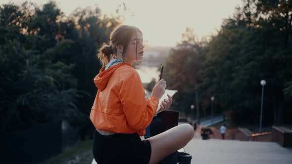 A Creative Young Woman in Glasses Makes Notes in the Notebook in Her Hand Sitting on the Bench alt