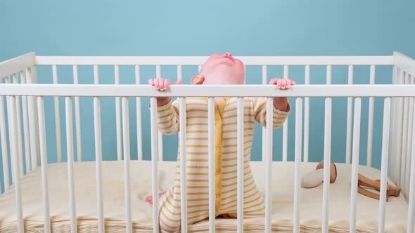 Cry infant baby boy stands in the crib, studio blue background. Sad child in yellow pajamas