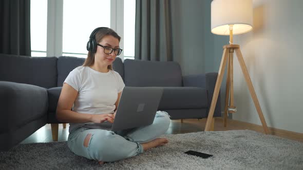 Casually Dressed Woman with Headphones is Sitting on Carpet with Laptop and Working in Cozy Room alt