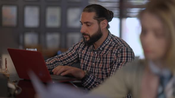Concentrated Man Texting Email on Laptop with Blurred Woman Analyzing Documents at Front alt