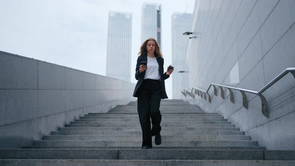 A Young Beautiful Unarmed Woman in a Business Suit Walks Down the Stairs Holding a Mobile Phone and alt