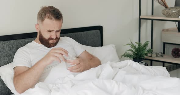 Young Bearded Guy Throwing Effervescent Cure Pill Into Glass of Water. Handsome Man in 30s Lying in alt