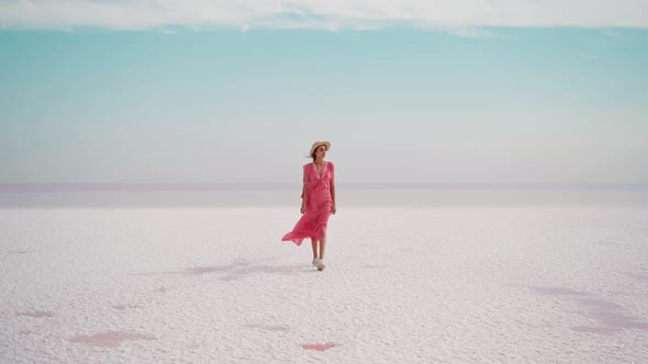 Carefree Elegant Woman in Blowing Pink Dress and Straw Hat Walking on White Salty Deserted Beach alt