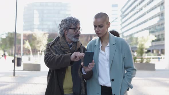 Two Business People Walking and Talking About Work While Checking Smart Phone alt