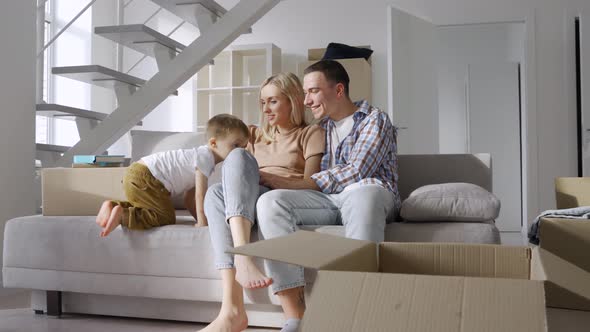 Tired but Happy Young Family with Boy Child Sitting at New Home at Moving Day alt