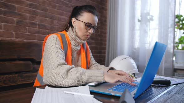 Woman Builder in Orange Helmet and an Orange Vest Working on a Laptop in the Room