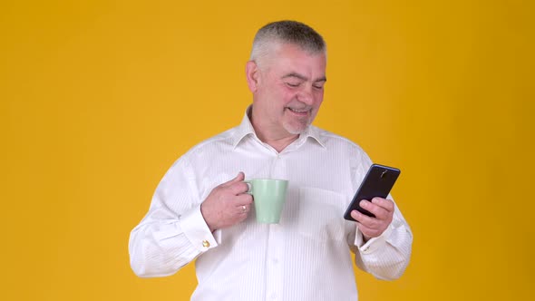 Positive adult man with a mug of coffee or tea uses smartphone texting messages in social media