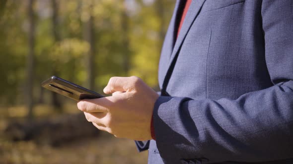 A Man Works on a Smartphone in a Park  Closeup  Trees in the Blurry Background