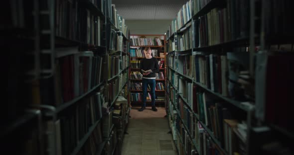 The Guy Is Standing Between the Bookshelves in the Light From the Window and Reading a Book. A alt