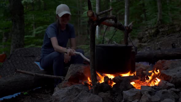 A Woman Watches the Fire While Cooking in Pot on a Campfire at Dusk