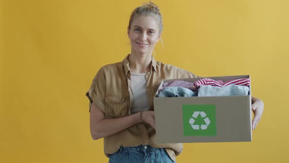 Slow Motion of Young Woman Holding Cardboard Box with Old Clothes for Recycling on Yellow Background alt