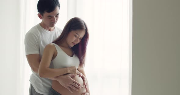 Asian husband with a pregnant wife at home in the bedroom holding her belly.