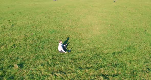 Low Orbital Flight Around Man on Green Grass with Notebook Pad at Yellow Rural Field. alt
