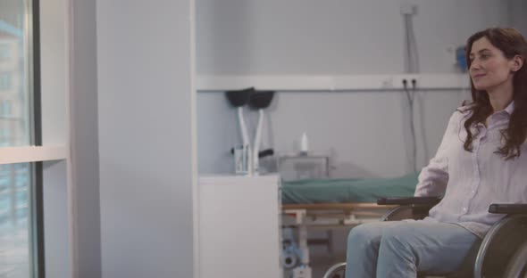 Smiling Female Patient Sitting in Wheelchair in Hospital Room Looking Through Window alt