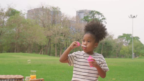 Happy little african american curly hair girl blowing soap bubbles playing alone in the park alt