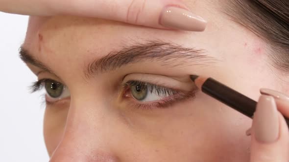 Professional Stylist Makeup Artist with Brown Pencil Draws Arrows in Eye Makeup to Young Beautiful alt