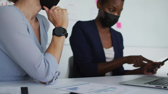 Two diverse female colleagues wearing face mask, sitting at desk and talking alt