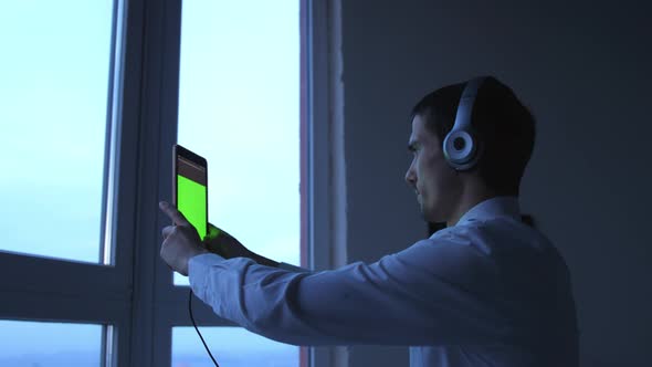 Young Businessman in Headphones Uses Tablet By the Window in the Evening