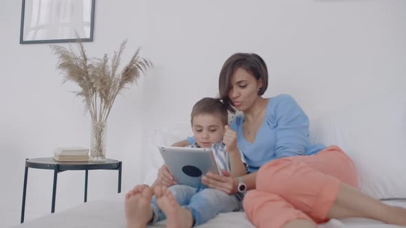 In the White Bedroom Mom and Son Look at the Tablet Screen and Laugh alt