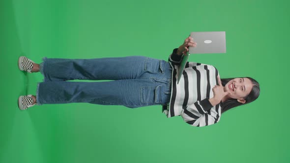 Full Body Of A Smiling Asian Woman Pointing To Computer While Standing In Front Of Green Screen alt